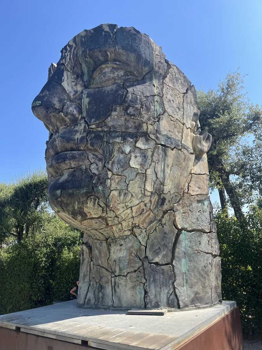 Distant portrait view of Mitoraj's Tindaro Screpolato at the Prato dei Castagni, Boboli Gardens, seen across the gravel path with the sculpture and its Corten plinth centred against a clear Florentine sky