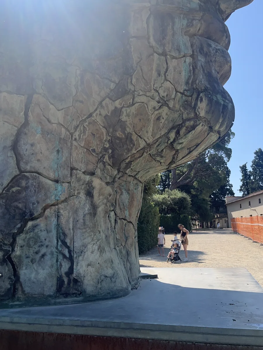 Wide low-angle view of the base of Tindaro Screpolato in the Boboli Gardens — the massive cracked bronze curving overhead as a young family with a pushchair walks beneath, umbrella pines beyond