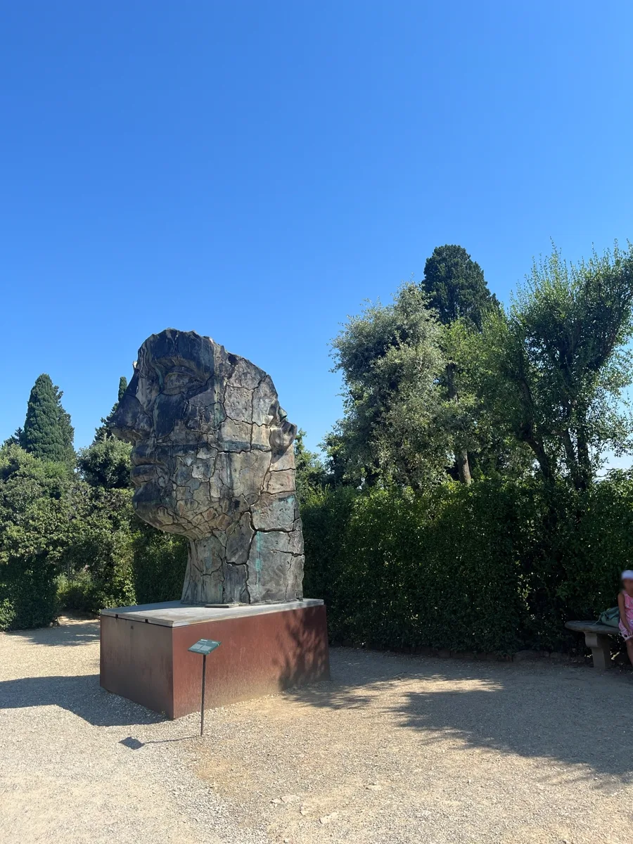 Distant three-quarter view of Igor Mitoraj's Tindaro Screpolato in the Boboli Gardens, framed by cypress and holm oak, a solitary visitor seated at right