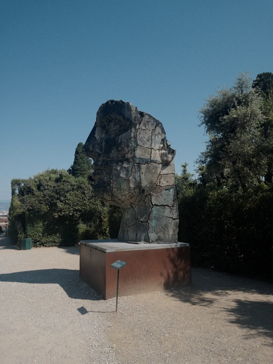 Wide landscape view of Igor Mitoraj's Tindaro Screpolato standing on its Corten plinth at the Prato dei Castagni, the Tuscan hills of Florence visible in the far distance