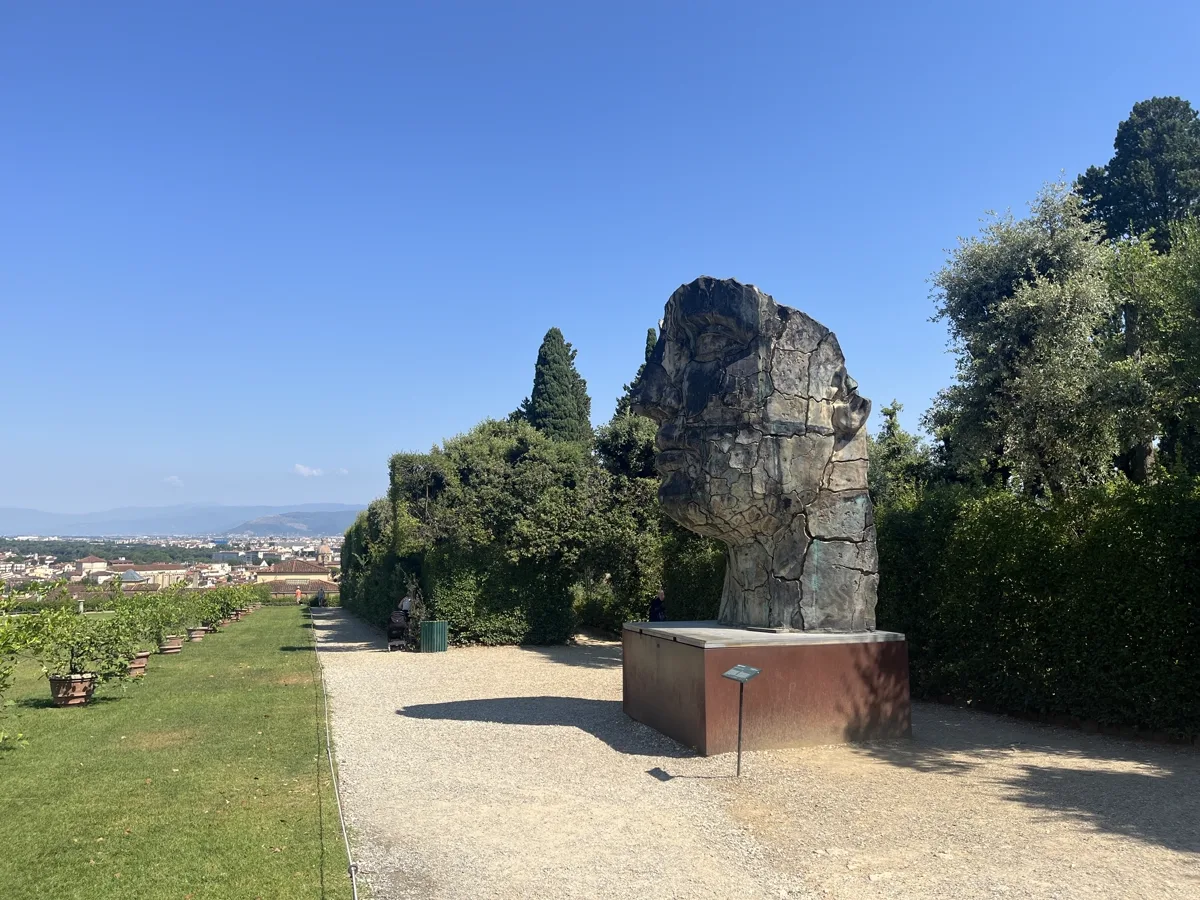 Broad landscape view showing Tindaro Screpolato at right and the Florentine hills to the left, the sculpture standing above trimmed hedges on the upper Boboli terrace