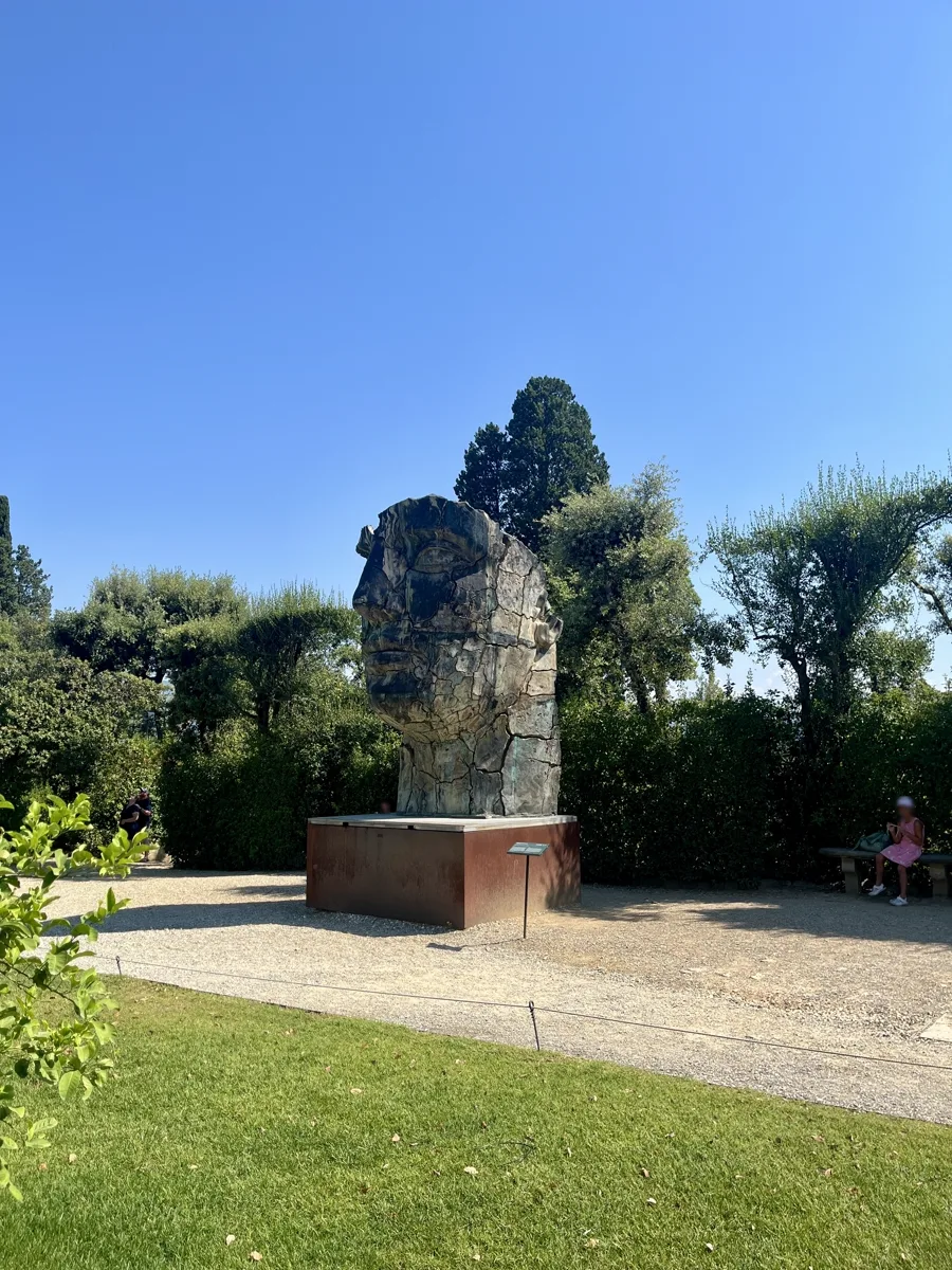 Tindaro Screpolato viewed across a stretch of Boboli lawn from a distance, the cracked bronze face framed by dense summer foliage and a low bench at the right edge