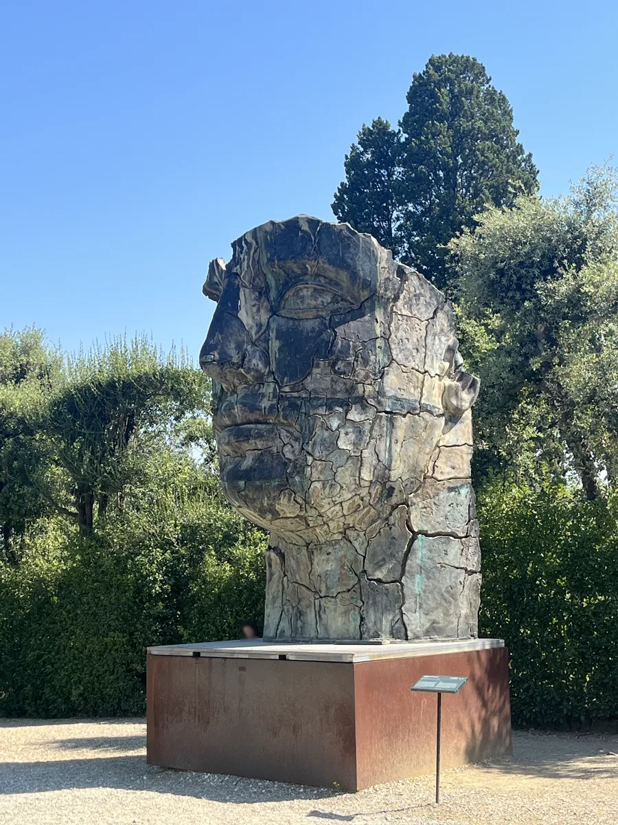 Front elevation of Igor Mitoraj's Tindaro Screpolato showing the full facial profile in sunlight, the bronze silhouetted between two dark cypresses in the Boboli Gardens