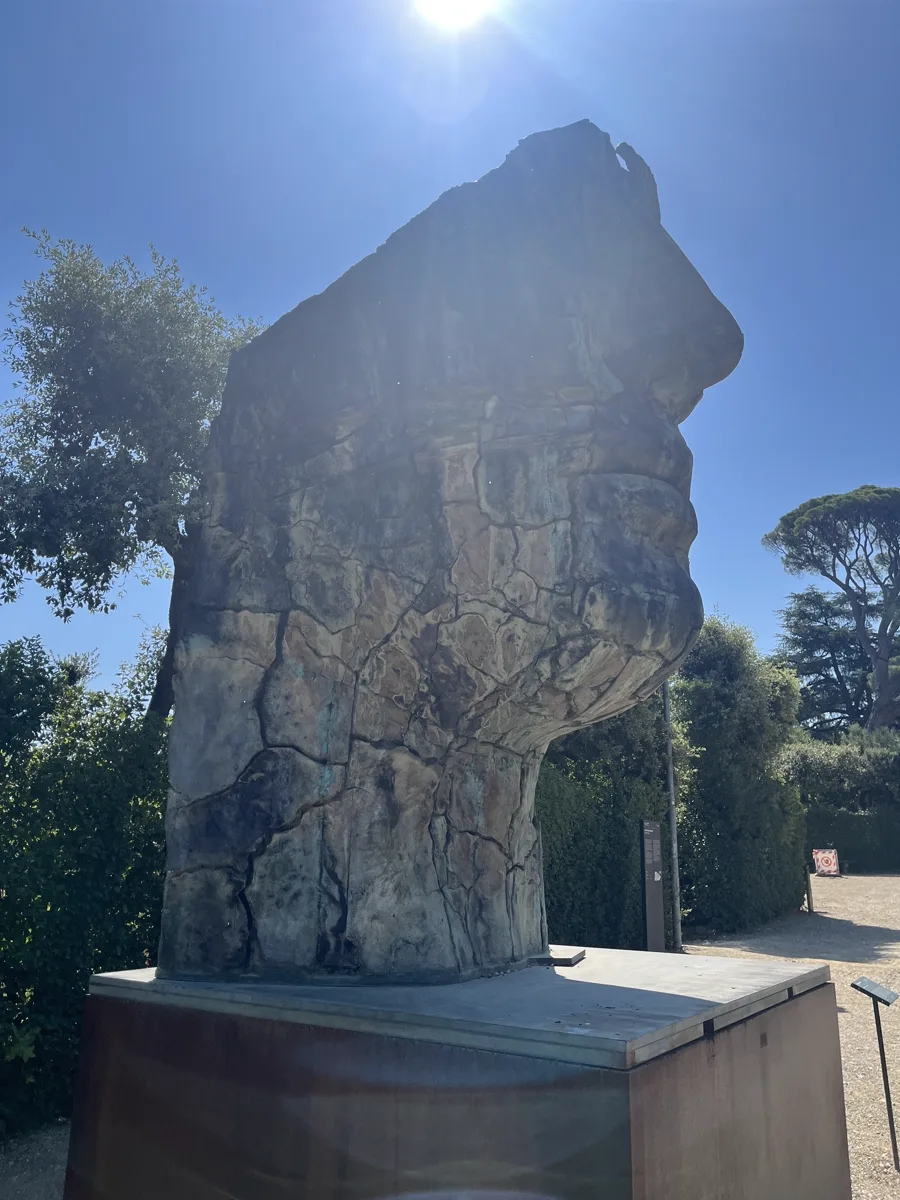 Another contre-jour view of Tindaro Screpolato with the sun almost directly above the sculpture, hedges and umbrella pines forming the darker frame around the bronze