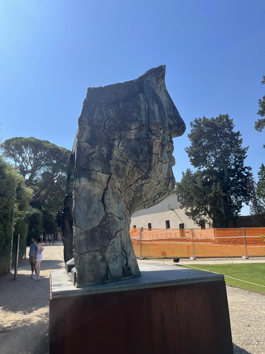Rear three-quarter view of Tindaro Screpolato in the Boboli Gardens with visitors passing along the walkway at left and an orange safety mesh marking a nearby garden works site