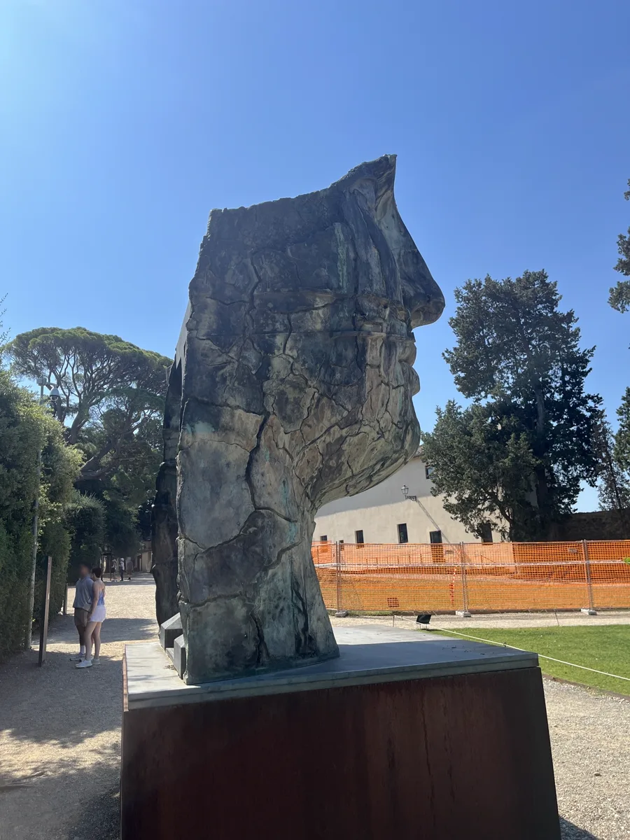 Back view of Tindaro Screpolato on its Corten plinth, the reverse of the cracked bronze mask facing the garden works fence, umbrella pines rising behind