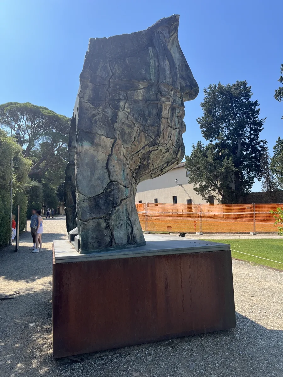 Rear elevation of Tindaro Screpolato photographed from further along the path, the back of the bronze reading as an abstract cracked mass against summer sky