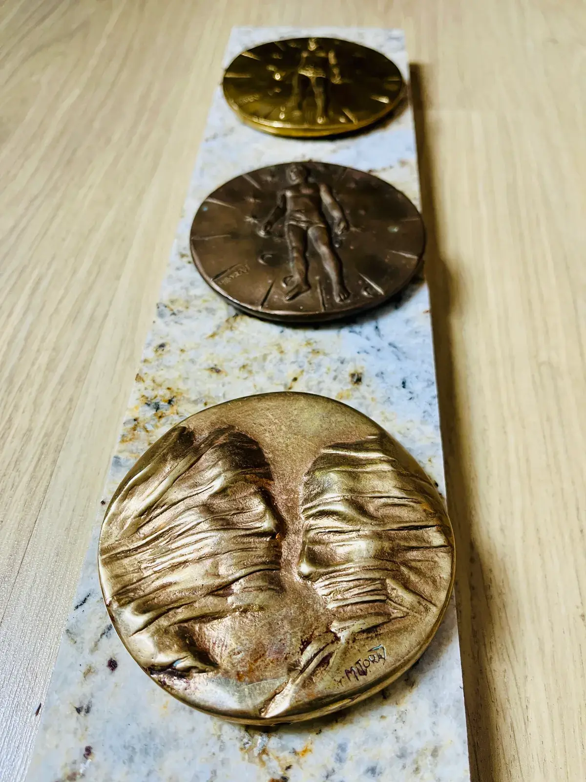La Conversation — Igor Mitoraj, three medals vertical on marble: gilt reverse (top), dark reverse (middle), worn obverse (bottom)